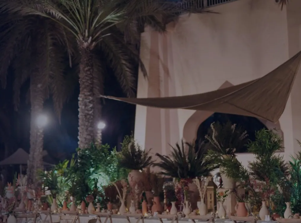 Palm trees above a long dinner table dressed with greenery and earthenware, lit at night.